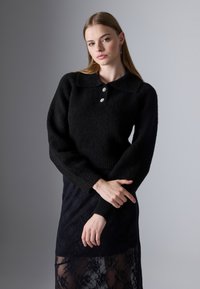 Pulover negru tricotat cu guler, având trei nasturi. Asortat cu o fustă neagră din dantelă, texturată și transparentă, care evidențiază detaliile de straturi.