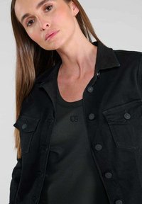 Zwarte denim jasje met een puntige kraag, knoopsluiting en twee borstzakken. Draag het over een donkergroene geribbelde tanktop. Gladde textuur.