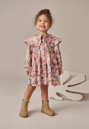 Lyserød blomsterkjole med lange ærmer, rynket krave og sammensyet nederdel. Brune ankelstøvler med dekorative syninger. Neutralt baggrund.