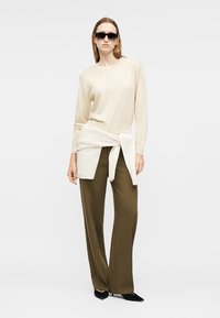 Femme portant un haut à manches longues beige, un pull crème noué autour de la taille, un pantalon large olive, des talons noirs pointus et des lunettes de soleil noires.