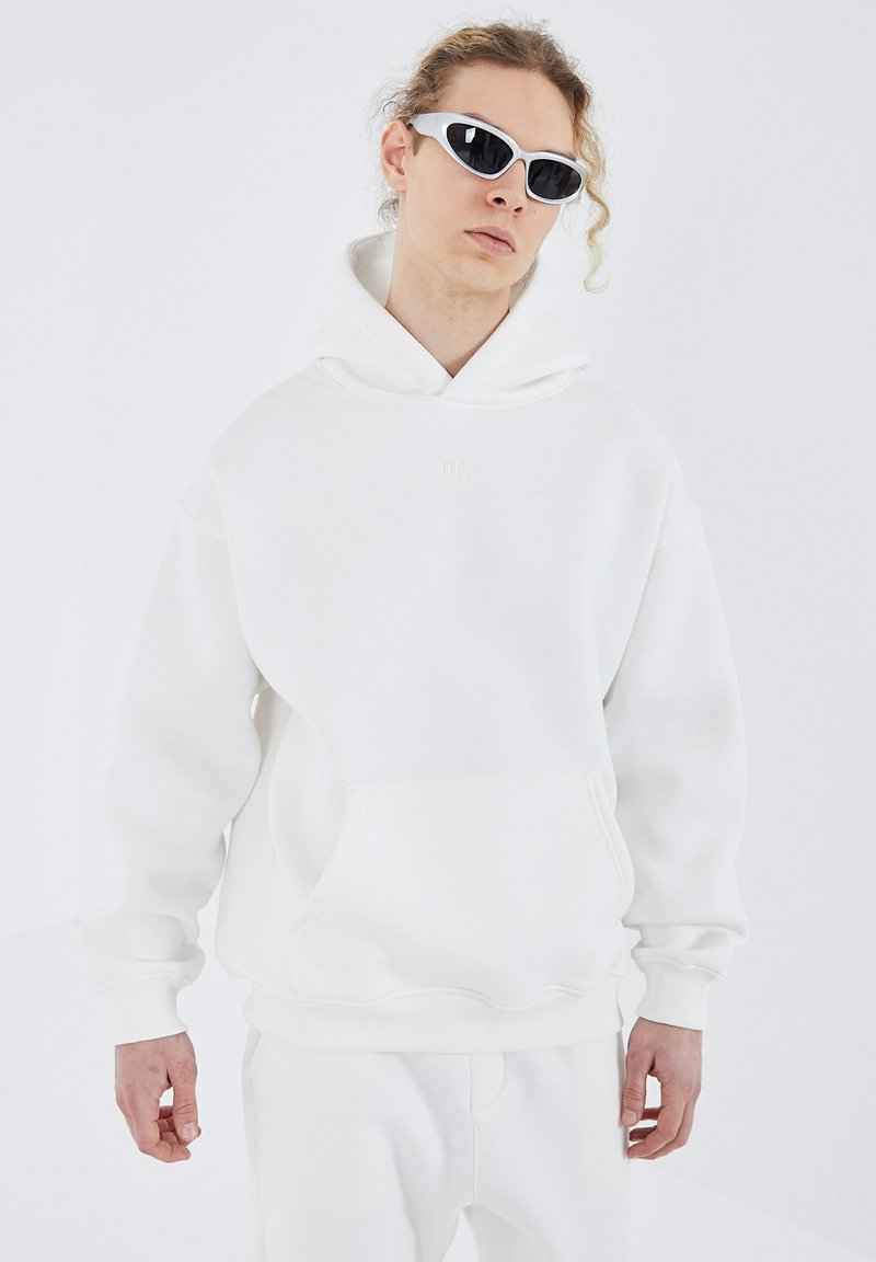 White hoodie with a kangaroo pocket, ribbed cuffs, and hem. Styled with sunglasses, featuring a relaxed fit and soft fabric texture.