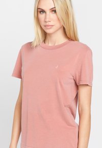 Rosa Baumwoll-T-Shirt mit kurzen Ärmeln, Rundhalsausschnitt und einem kleinen weißen Logo auf der linken Brust. Glatte Textur, entspannte Passform.