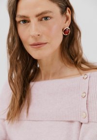 Světle růžový svetr s žebrovaným vzorem, který má široký výstřih a zlaté knoflíky na rameni. Texturovaná, měkká pletenina.