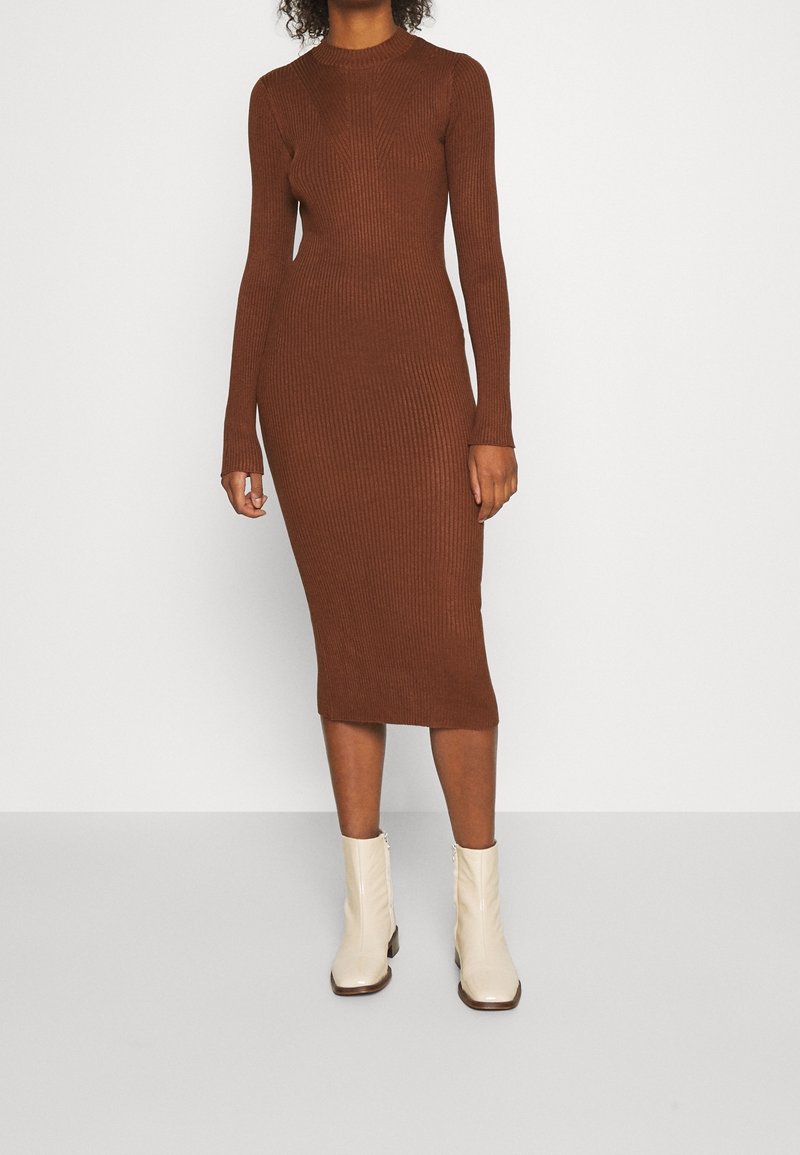 Femme portant une robe midi marron moulante à manches longues côtelées, associée à des bottines crème, se tenant devant un fond clair uni.