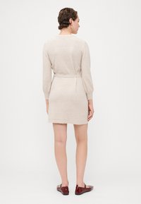 Beige strikket kjole med lange ermer, belte i midjen og rund hals, med en myk tekstur og en tettsittende silhuett, iført røde sko.