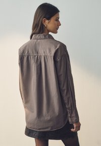 Brown, oversized shirt made of soft cotton with a classic collar, buttoned cuffs, and pleated detailing on the back. Simple design.