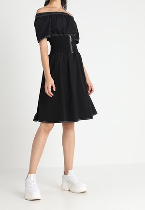 Vestito nero con spalle scoperte, vita arricciata e dettaglio con cerniera, caratterizzato da una gonna svasata, abbinato a sneaker bianche chunky.