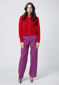 Cardigan rosso a maglia con bottoni, abbinato a pantaloni a gamba larga viola a vita alta e tacchi con punta in argento. Tessuti lisci e vestibilità su misura.