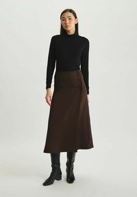 Jupe portefeuille marron de longueur genou avec des boutons sur le side, fabriquée en tissu lisse. Associée à un haut noir à manches longues et des bottes noires montantes.