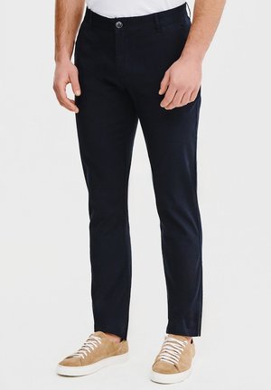 Granatowe, slim-fit chinos z płaskim przodem i zapięciem na guzik; w połączeniu z beżowymi sneakersami z białymi sznurowadłami i teksturowaną powierzchnią.
