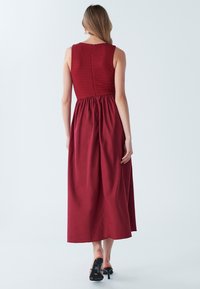 Rdeča maksi obleka z oprijetim bodijem iz nabranega blaga in ohlapno krilom. Ima okrogel izrez, zadrgo na zadnji strani in rahlo tekočo teksturo tkanine.