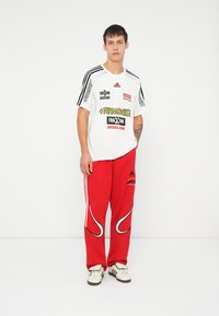 Bílá sportovní tričko s černými a červenými detaily, s různými logy. Červené tepláky s bílým bočním lemováním a logem Adidas.