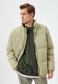 Lysegrøn pufferjakke med høj krave og lynlås, båret over en mørkegrøn sweatshirt. Ribmede manchetter og kant, glat tekstur.