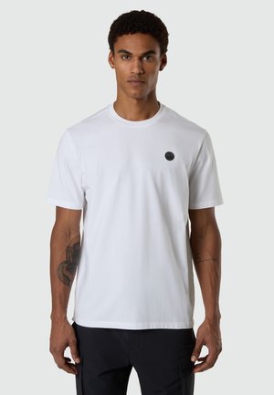 Giovane con capelli ricci corti che indossa una semplice t-shirt bianca a maniche corte con un piccolo logo nero circolare sul petto.