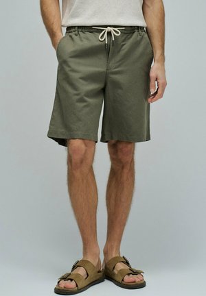 Hombre con pantalones cortos verde oliva con cordón y sandalias marrones con hebilla, de pie con las manos en los bolsillos frente a un fondo liso.