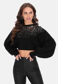 Fekete, rövidített pulóver, oversized ujjakkal, amelyet fényes fém flitterek díszítenek a nyakrészen. Magas derekú fekete nadrággal párosítva.