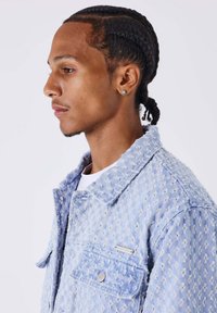 Lichtblauwe spijkerjasje met een gestructureerd patroon, knoopzakken en een kraag. Gedragen over een wit shirt, met een zilveren accenten logo.