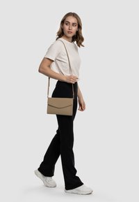 Borsa a tracolla beige con superficie texture, forma rettangolare e chiusura a patta, abbinata a pantaloni neri e sneakers bianche.
