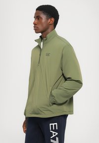 Grønn zip-up jakke med høy krage, lett stoff, subtil logo på brystet og elastiske mansjetter. Kombinert med marineblå bukser med hvit tekst.