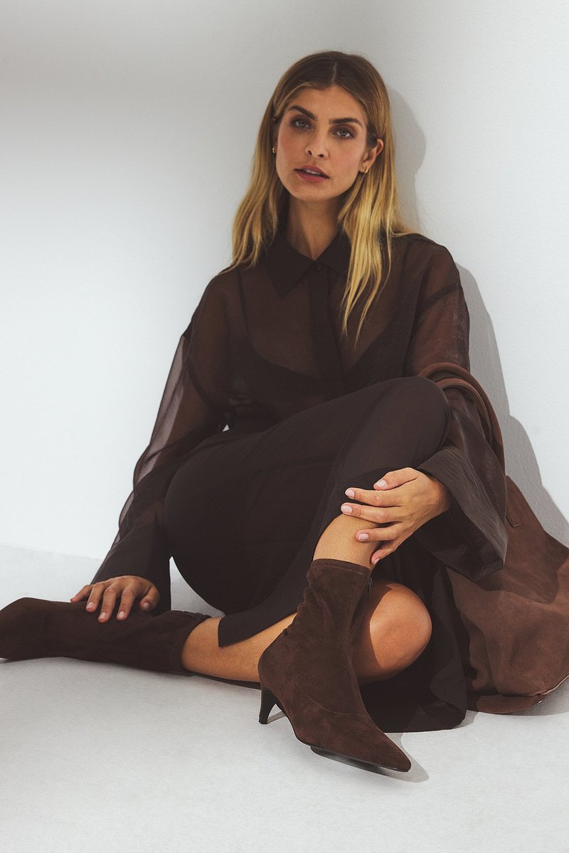 Bottes de cheville en suède brun avec un bout pointu et un talon fin, assorties avec une tenue fluide et transparente de couleur brun foncé et un châle texturé.