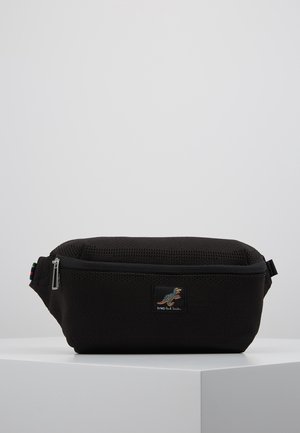 Schwarze Stoff-Gürteltasche mit Reißverschluss, strukturierter Oberfläche und kleinem buntem Dinosaurier-Patch vorne, auf weißem Podest vor hellgrauem Hintergrund platziert.