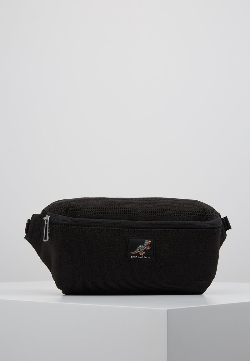 Sac banane en tissu noir avec fermeture éclair, surface texturée et petit patch dinosaure coloré à l'avant, posé sur un socle blanc sur fond gris clair.