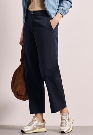 Personne portant un pantalon court bleu marine, des baskets beiges et une veste bleu clair, tenant un grand sac en daim marron.