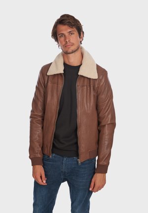 Chaqueta bomber de cuero marrón con un cuello de forro polar color crema, puños acanalados y dobladillo. Combinada con una camisa negra y jeans azules.