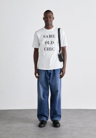 White T-shirt with black text "SAME OLD CHIC," paired with loose blue denim jeans, and black sneakers. Crossbody bag attached.