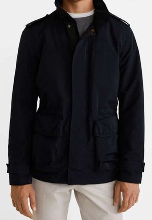 Mann trägt eine schwarze Kapuzenjacke mit aufgesetzten Fronttaschen und knöpfbaren Bündchen, kombiniert mit beigen Hosen.