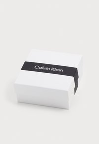 Fehér négyzet alakú doboz fekete sávval, amelyen a "Calvin Klein" felirat található. Simaszerkezet, minimalista dizájn és látható hardver nélkül.