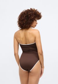 Brūns ribots viengabala peldkostīms ar baltām akcentiem, kuram ir bezsiksnas dizains un vidēji augsta kāja. Gluda tekstūra un pieguļošs siluets.