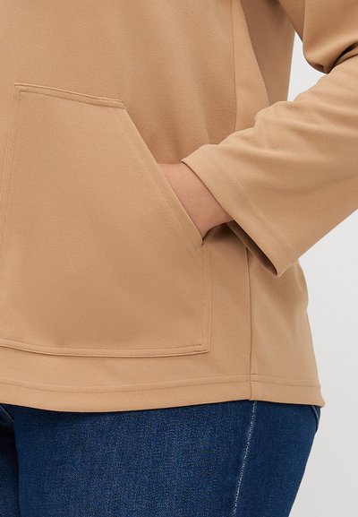 Pull beige à manches longues avec une poche avant. Texture douce et design décontracté, assorti à un jean en denim foncé.