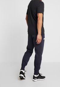 Man som står vänd åt sidan och bär en svart t-shirt, marinblå joggers med bakfickor, vita strumpor och svarta Under Armour-sneakers.
