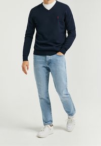 Pull col V bleu marine en tissu tricoté, associé à un jean denim bleu clair, des baskets blanches et un t-shirt blanc en dessous.