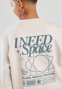 Beige sweatshirt featuring a large blue graphic text "I NEED Space" with stars, an atom design, and a barcode. Soft, textured fabric.