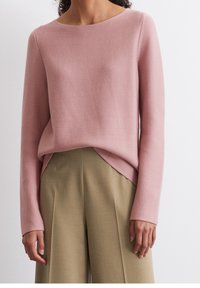 Femme portant un pull en maille rose poudré avec un ourlet asymétrique, rentré dans un pantalon taupe taille haute à jambes larges.