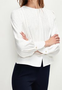 Witte blouse met lange mouwen, voorzien van een gestructureerd bloemenpatroon, plooien aan de voorkant en knoopmanchetten. Draag een navy broek.