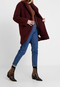 Cappotto in pelliccia sintetica borgogna con texture, colletto e apertura frontale; abbinato a jeans blu a vita alta e stivaletti alla caviglia con stampa serpente.