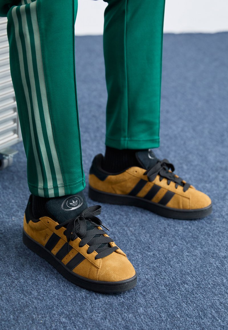 Brown suede sneaker with black accents, three black stripes, and a black rubber sole, paired with green athletic pants featuring white stripes.