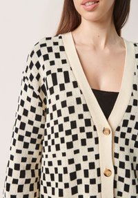 Svart och krämfärgad rutmönstrad cardigan med v-ringning, knäppning och stickad textur. Märkbara träknappar ger detaljer till designen.