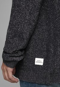 Gros plan sur la manche et le bas du dos d'un pull en maille gris foncé, avec une étiquette rectangulaire blanche Jack & Jones Originals au-dessus d'un jean bleu.