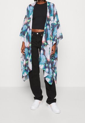 Person, die ein buntes, floral gemustertes, offenes Kimono, ein schwarzes Crop-Top, schwarze hoch taillierte Hosen und weiße Sneaker trägt, steht vor einem einfarbigen Hintergrund.