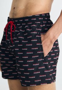 Șorturi de înot negre cu talie elastică roșie și șnur, prezentând un model repetat cu textul "HUGO" în roșu și alb. Include un buzunar lateral.