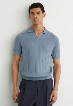 Uomo che indossa una polo lavorata a maglia azzurro chiaro a maniche corte con righe verticali, in piedi contro un muro bianco, abbinata a pantaloni scuri.