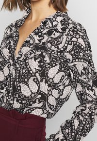 Schwarze Bluse mit weißem floralem Paisley-Muster; verfügt über einen Kragen, eine geknöpfte Leiste und lange Ärmel, aus einem leichten, strukturierten Stoff gefertigt.