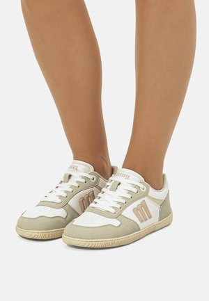Gros plan d'une personne portant des baskets lacées blanches et beiges avec des semelles beiges, debout sur un fond blanc.