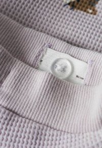 Vêtement en coton lilas avec une surface texturée, doté d'une patte de boutonnage blanche lisse et d'un bouton transparent, affichant des détails de couture soignés.