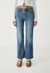 Vaqueros de campana de denim azul claro con un aspecto desgastado, que cuentan con cinco bolsillos y un cierre de botón, combinados con zapatillas marrones.