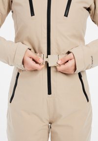 Completo beige con dettagli neri e zip; le mani serrano una fibbia chiara in vita. Presenta maniche lunghe e tasche.